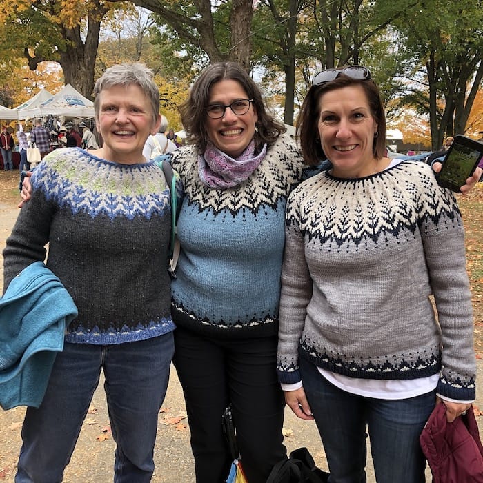 Three women stand close, wearing coordinating stranded circular yoke sweaters in shades of grey, blue, and cream.