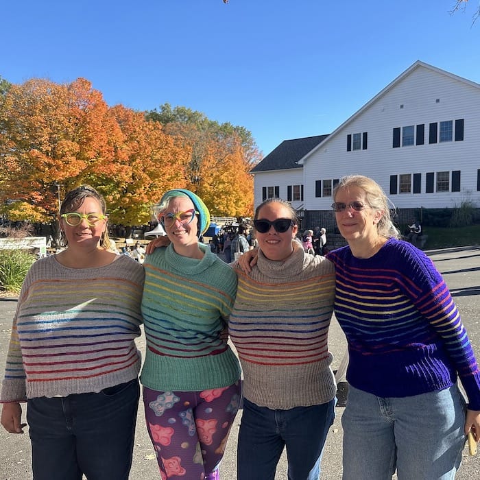 Four smiling women in matching sweaters, all in solid colors with thin rainbow stripes.