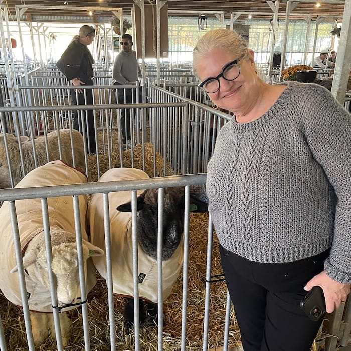 A woman stands smiling next to two sheep, wearing a grey crocheted sweater in a gansey textured pattern.