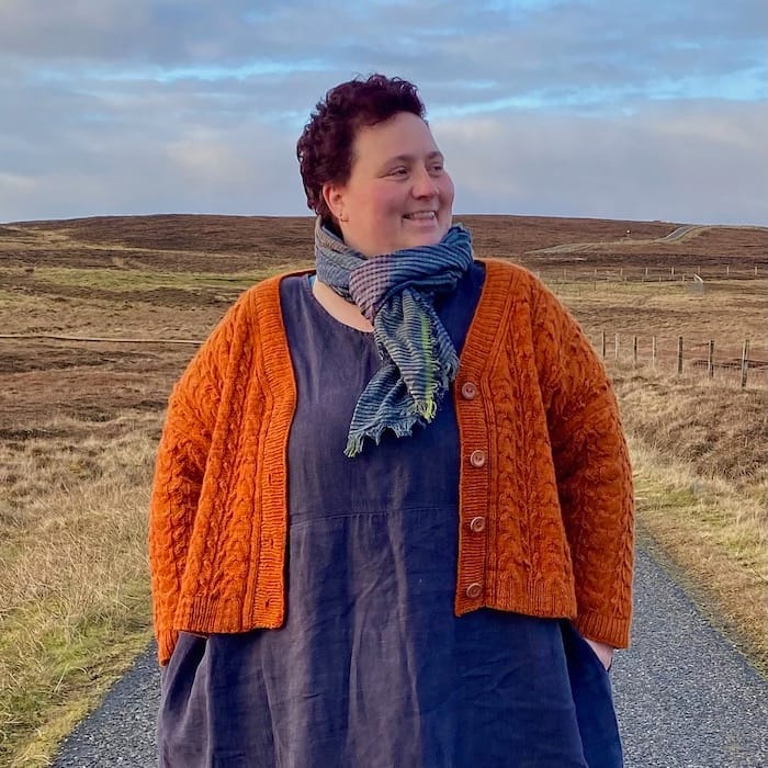 Beaming against a spacious sky and field, a woman wears a rich orange cabled cardigan with yarn from her own yarn line.