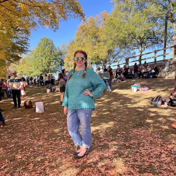 A woman stands on a hill in the shade of an autumn tree, wearing an aqua sweater and jeans. She is smiling happily.