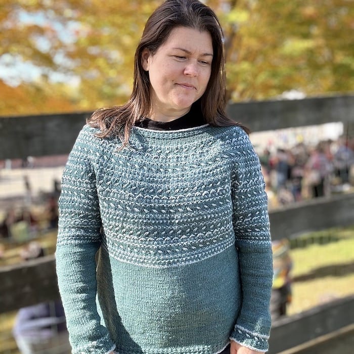 A woman smiles softly, looking down, wearing a warm aqua circular yoked sweater with textured white stranded colorwork.