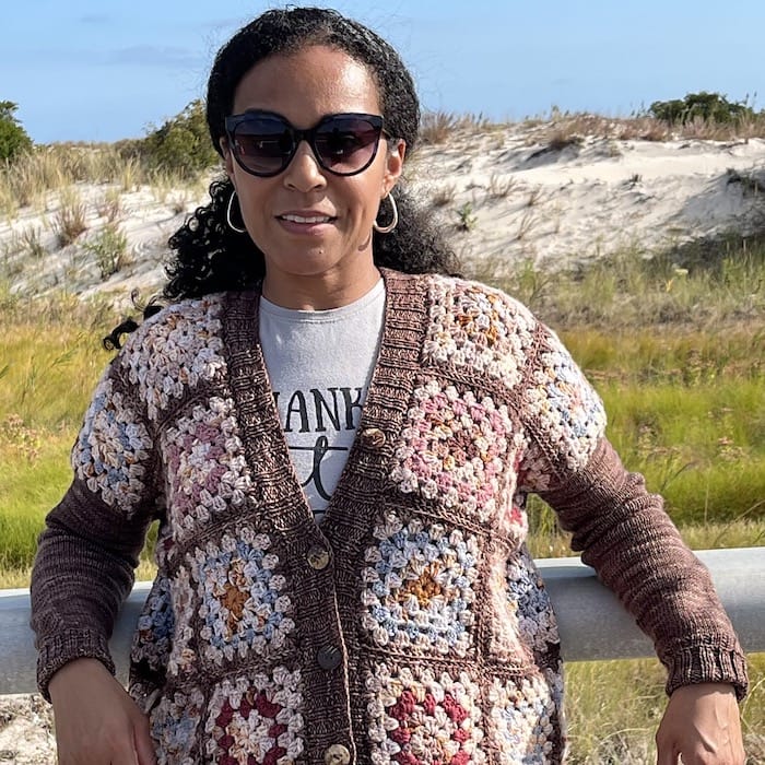 A woman in a crocheted granny squares cardigan made in soft neutrals stands on a beachy path, smiling at the camera.