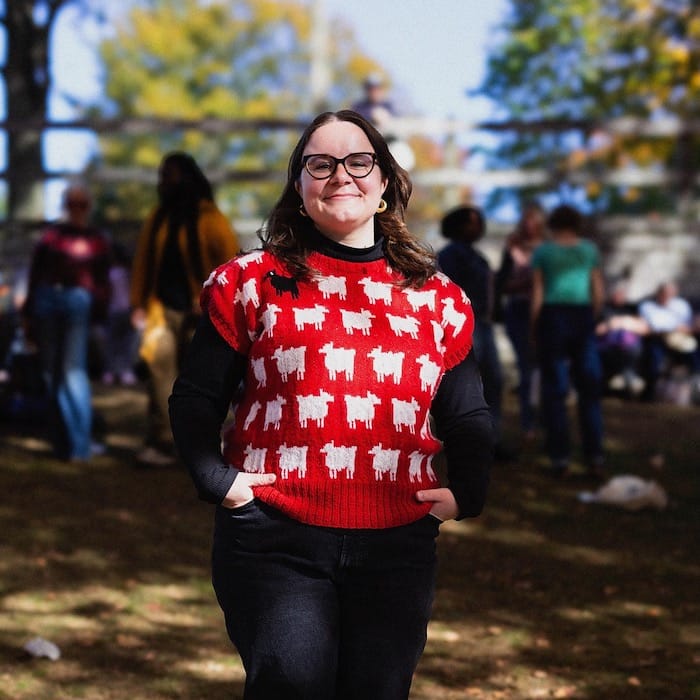 A woman stands proudly in dappled sunlight, wearing a vest that replicates Princess Diana's famous "black sheep" sweater.