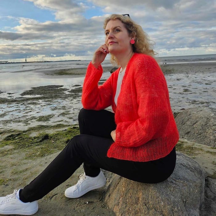 Against a beautiful beach sunset, a woman sits on a rock with her head on her hand wearing a vibrant tomato red cardigan.