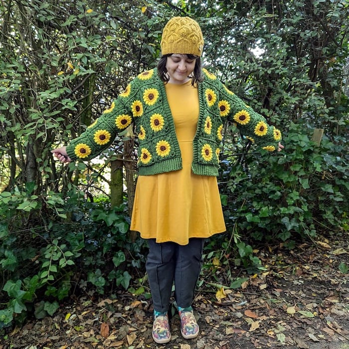 A woman stands in the woods showing off her whimsical crocheted cardigan, made from granny squares with sunflower centers.