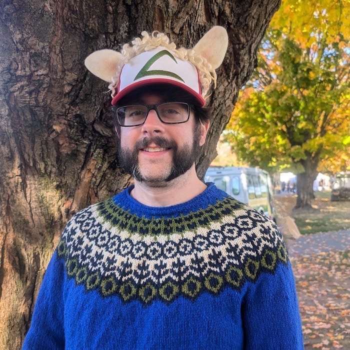 A man stands by a tree, smiling in a colorwork yoke sweater with a rich royal blue main color and colorwork in navy, olive green, and tan.