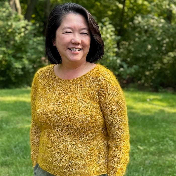 A woman in a yellow feather-and-fan sweater with a soft yarn halo smiles in a wooded clearing.