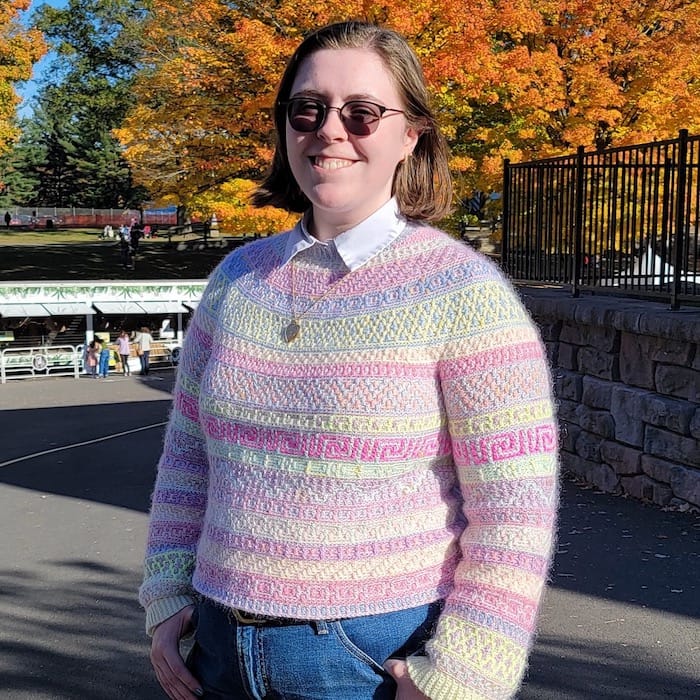 A woman stands on a pathway, against a backdrop of autumn leaves, smiling. She wears a crocheted mosaic sweater in shades of pink, cream, and lavender, with a soft halo.