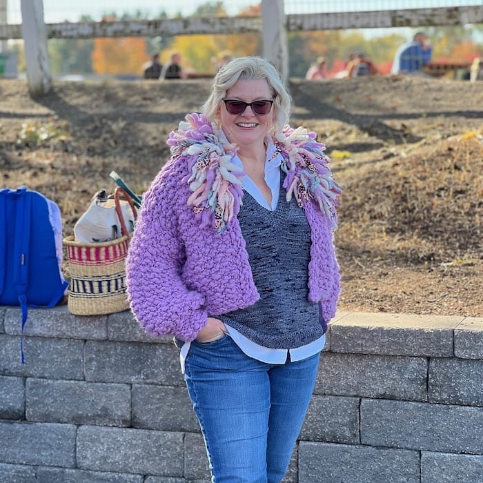 A woman stands against a stone wall wearing a textured chunky lilac cardigan with a dramatic fringe collar.