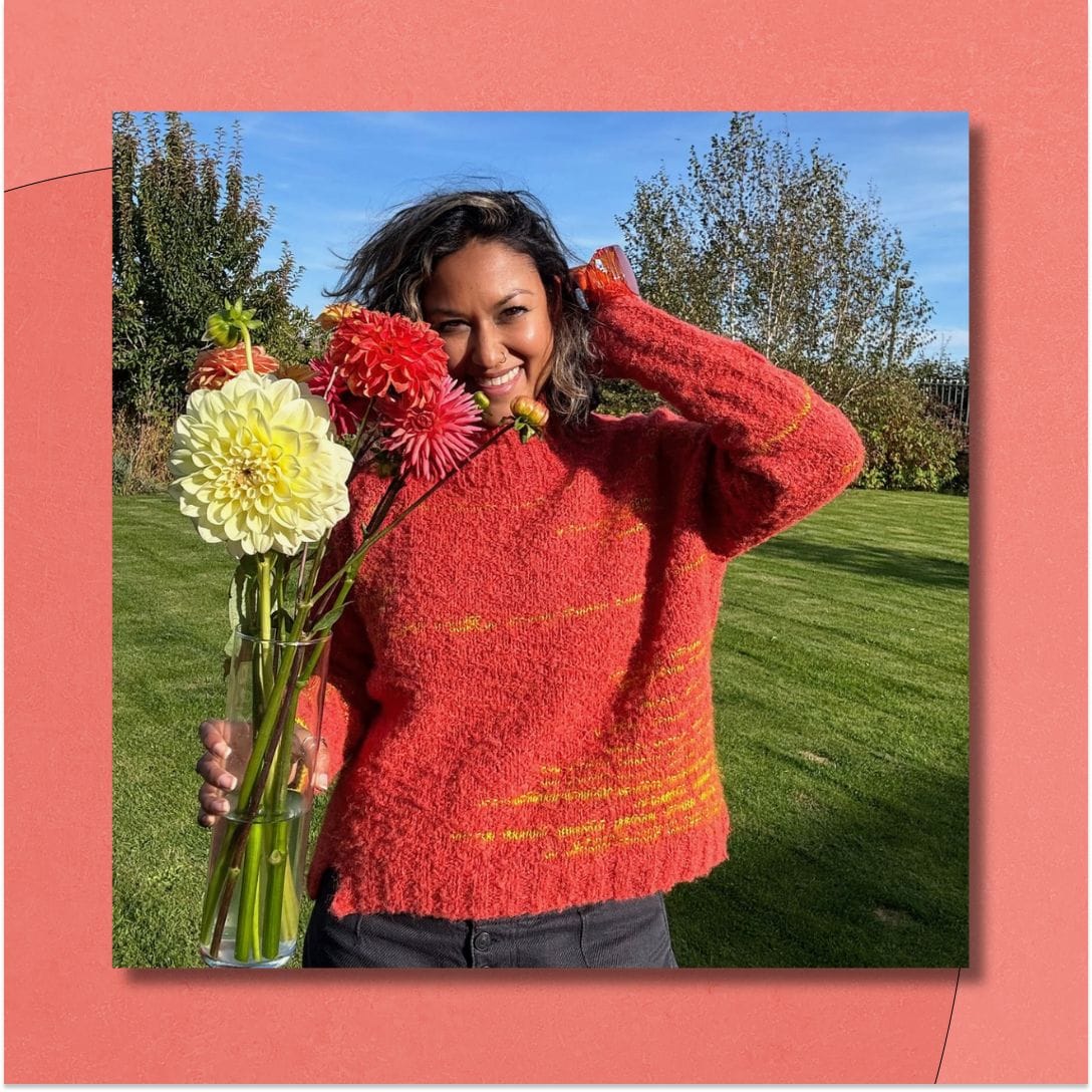 A smiling woman stands in a field with trees, holding a vase of flowers. She wears a bright orange and yellow boucle sweater.