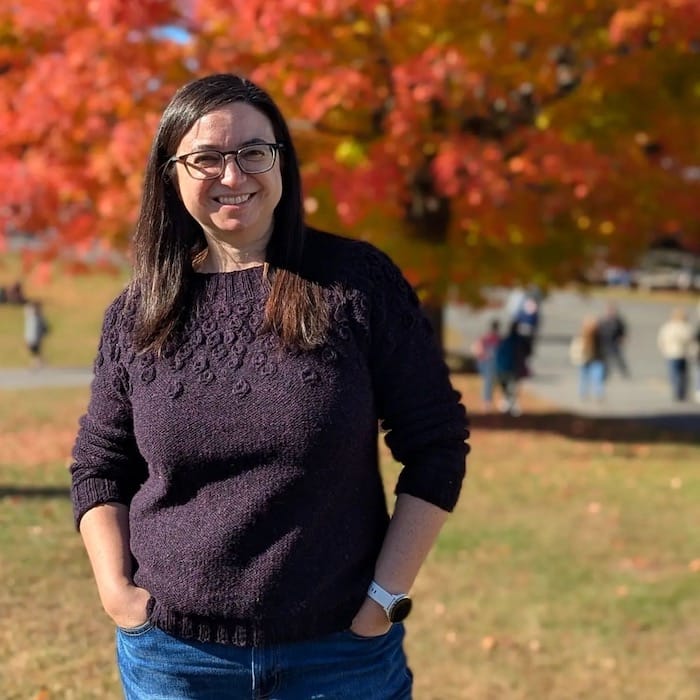 A woman stands, hands in jeans pockets, smiling against a backdrop of red autumn leaves. She is wearing a deep plum handknit sweater with a textured yoke.