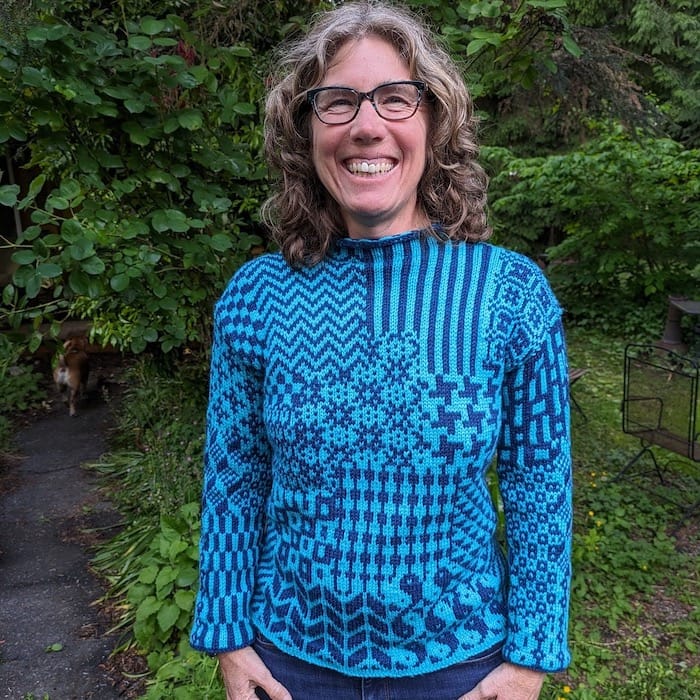 A woman stands facing the camera, beaming with joy in her stranded colorwork sweater, knit in blue and turquoise.