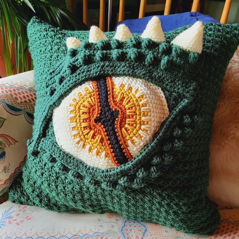 A bold, dramatic crocheted pillow made to look like a dragon's eye. The eye is gradient colors of orange, and the body of the pillow is a dark green. Above the eye are white dimensional spikes.