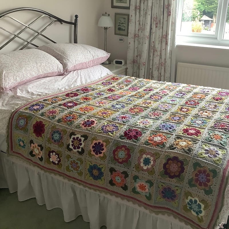 a bed is in the corner of a softly lit room, covered in a crocheted afghan made up of granny squares with center flowers, in vintage neutrals and pinks