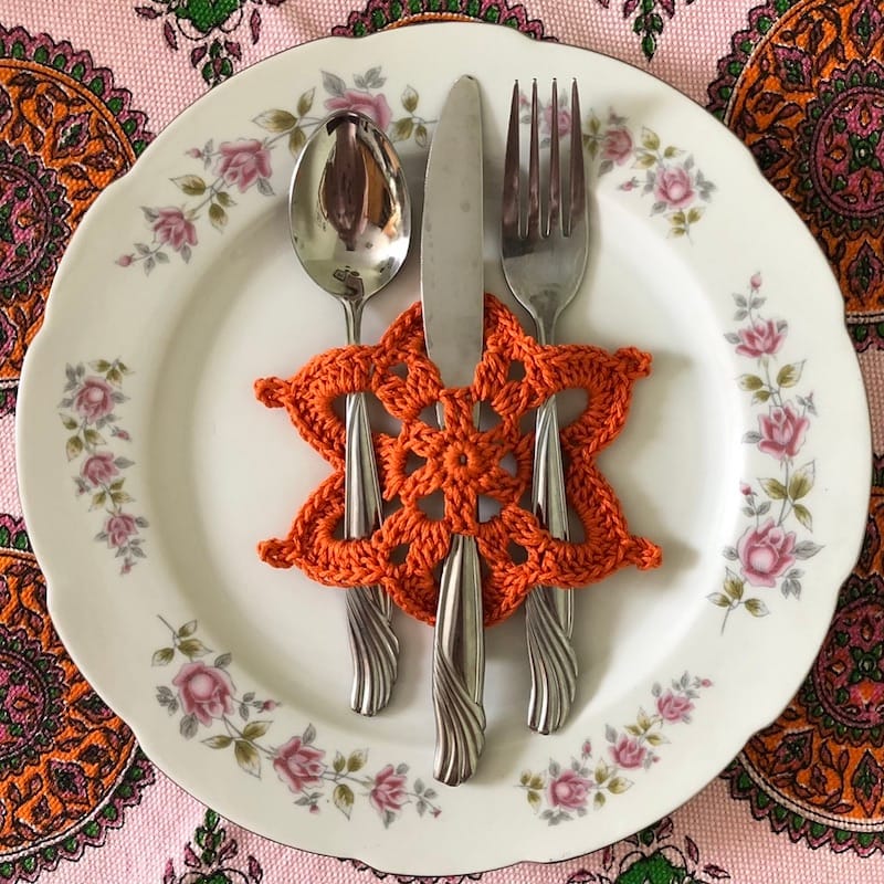 A delicate porcelain dinner plate with a silver place setting. The cutlery is woven through an orange crocheted snowflake motif.