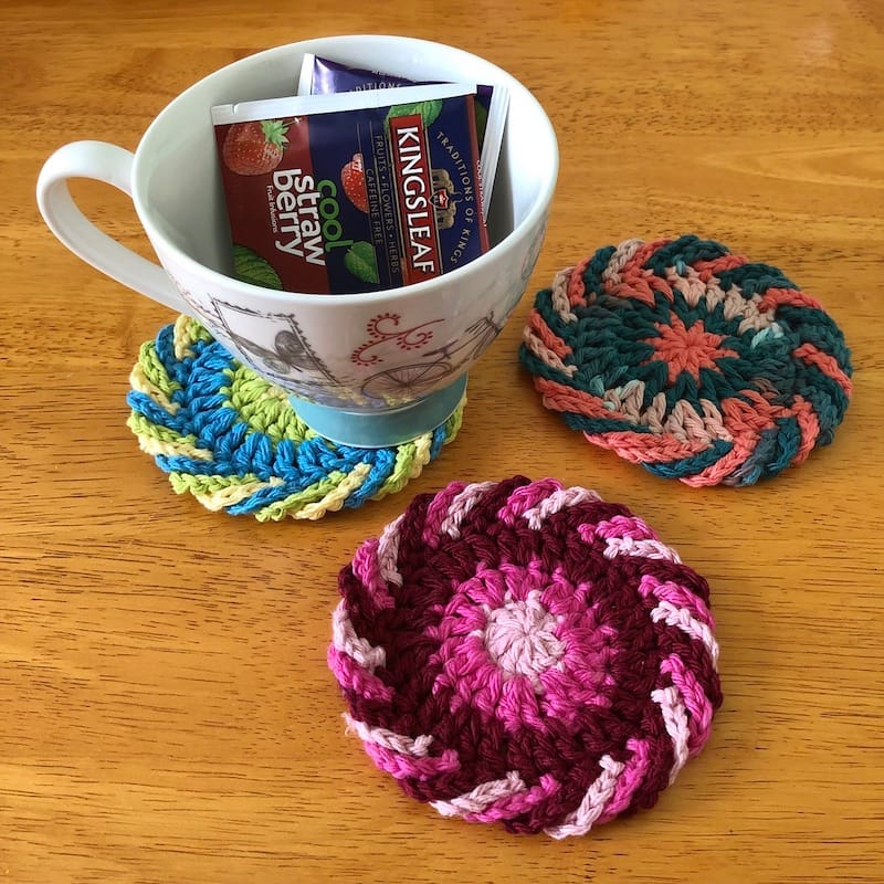 A trio of crocheted coasters in bright, cheery colors, on a table. A teacup ready with a bag of tea is atop one of them.