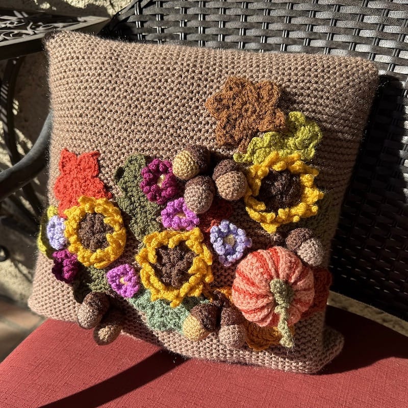 A whimsical crocheted pillow on an outdoor chair; it is a medium tan and adorned with crocheted flowers, acorns, leaves, and pumpkins.