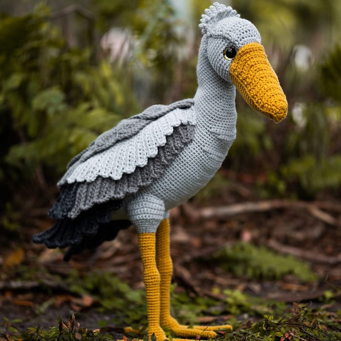 a regal crocheted shoebill stork stands in lush green nature. it is very detailed to look like the real creatures, with layers to its wings and a large bill.