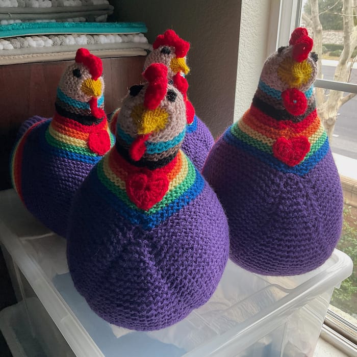 four knit chickens, mostly in purple but with neck stripes in Progress Pride flag colors, are perched near a window facing the camera