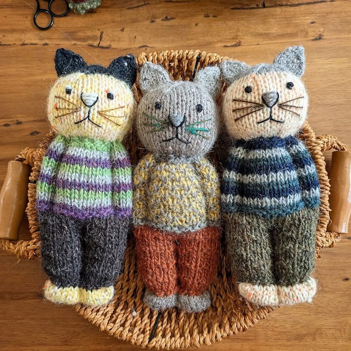 three knit cats are arranged in a basket next to each other. they are made to look as though they are standing like people, dressed in clothes.