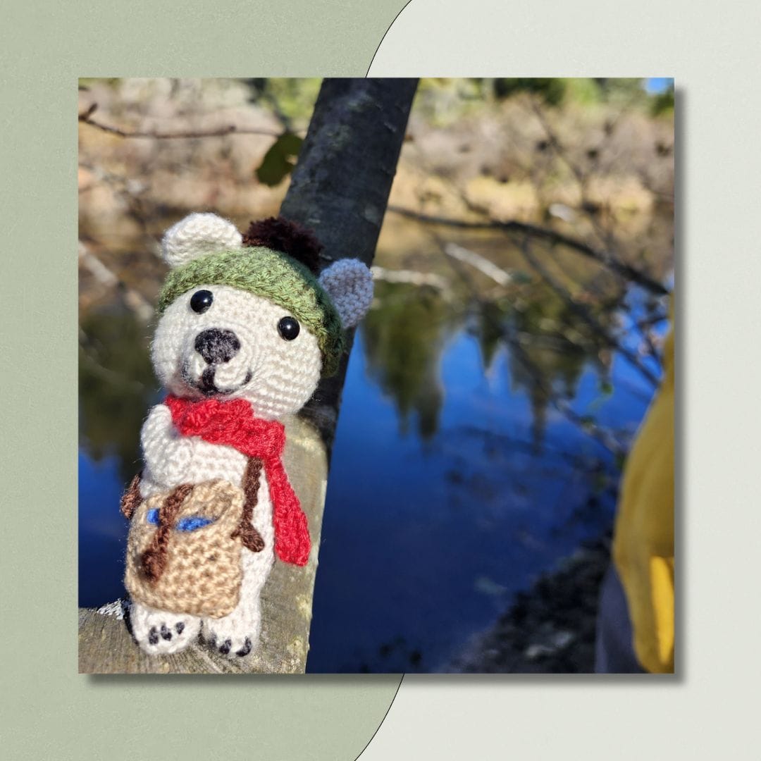 a charming crocheted teddy bear in a little green cap and red scarf holds a basket. the bear stands on a branch by a river.