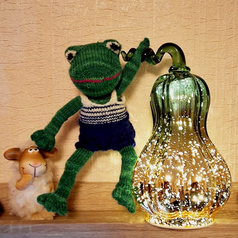 a handknit plush frog is posed jauntily on a shelf along with a sheep figure and glowing gourd