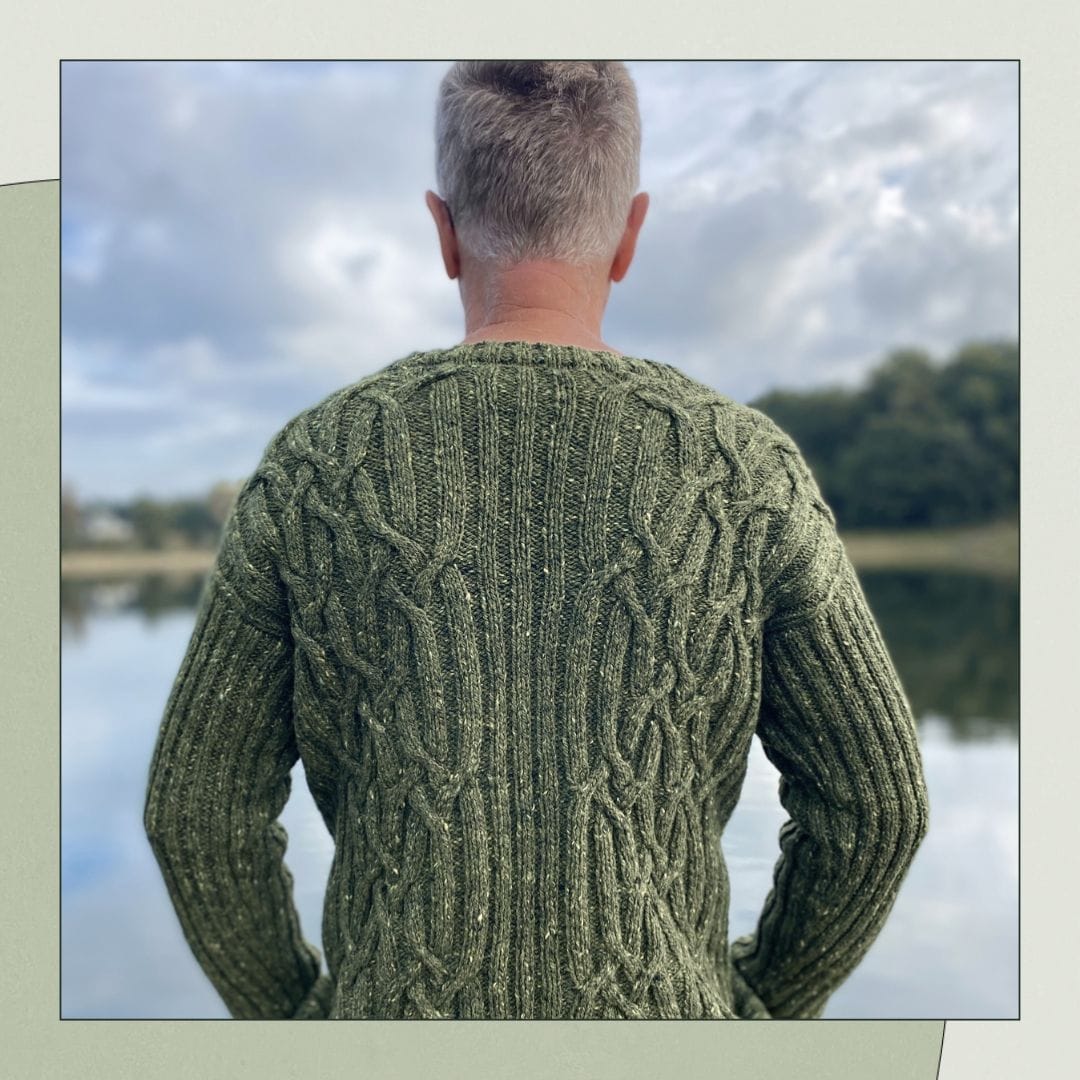 A person stands facing away from camera, looking toward a lake. They are wearing a cabled sweater in a tweed olive-green yarn.