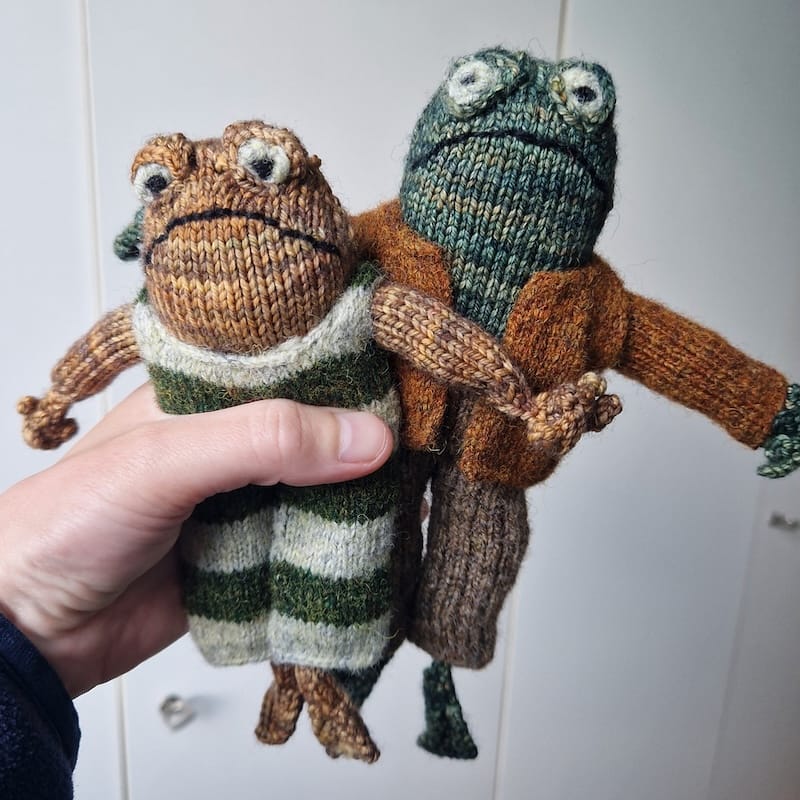 a hand holds up small knit stuffed plush figures of "Frog and Toad" from the book series