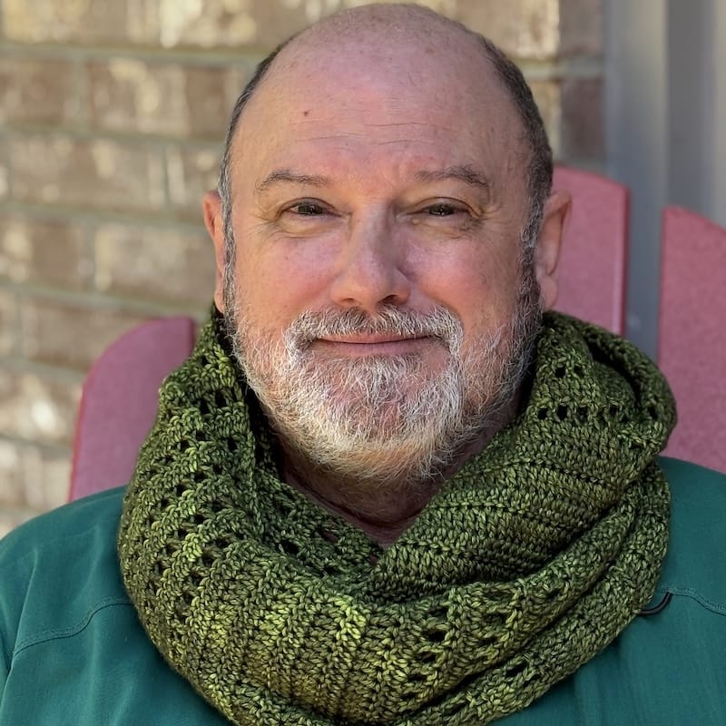a person looks at the camera with a glint of smile in their eyes, wearing an olive green slightly variegated crocheted wrap looped around their neck