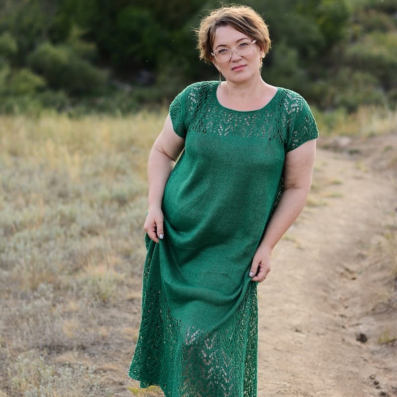 a person stands on a trail against a background of trees, looking at the camera, wearing a drapey green dress with lace details on the yoke, sleeve, and skirt