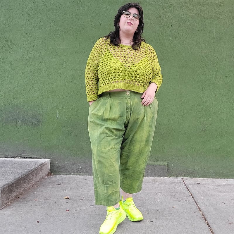 standing confidently facing the camera against a green backdrop, a person wears a handknit openwork sweater in a bright green, along with green pants and sneakers