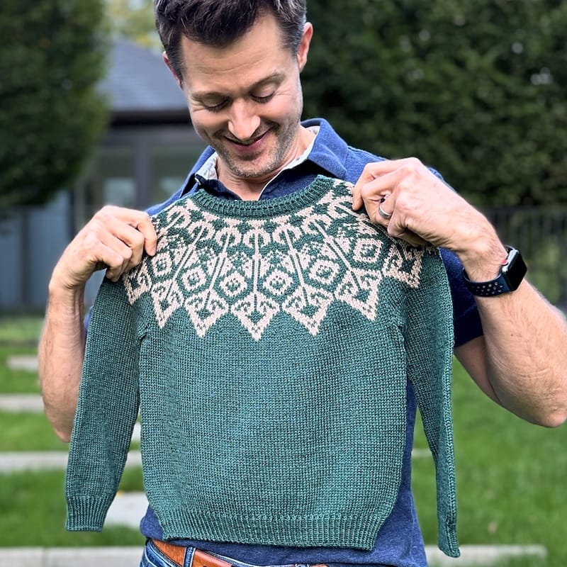 a person stands holding a completed green sweater with a colorwork yoke to their chest, smiling