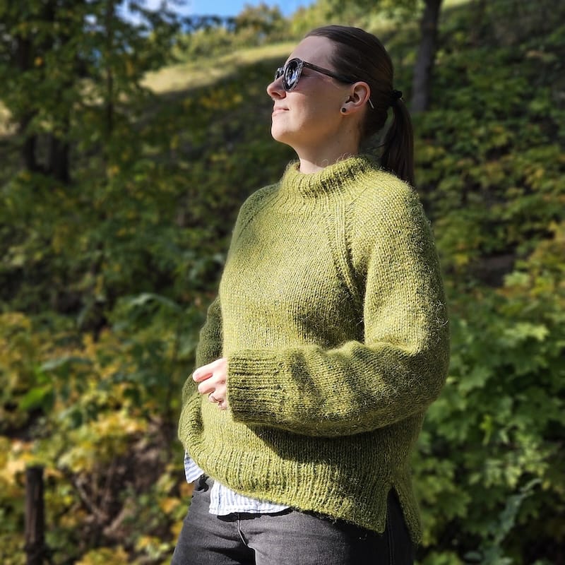 a person stands with their face toward the sun, filtered through leaves, wearing a cozy and oversized green knit sweater